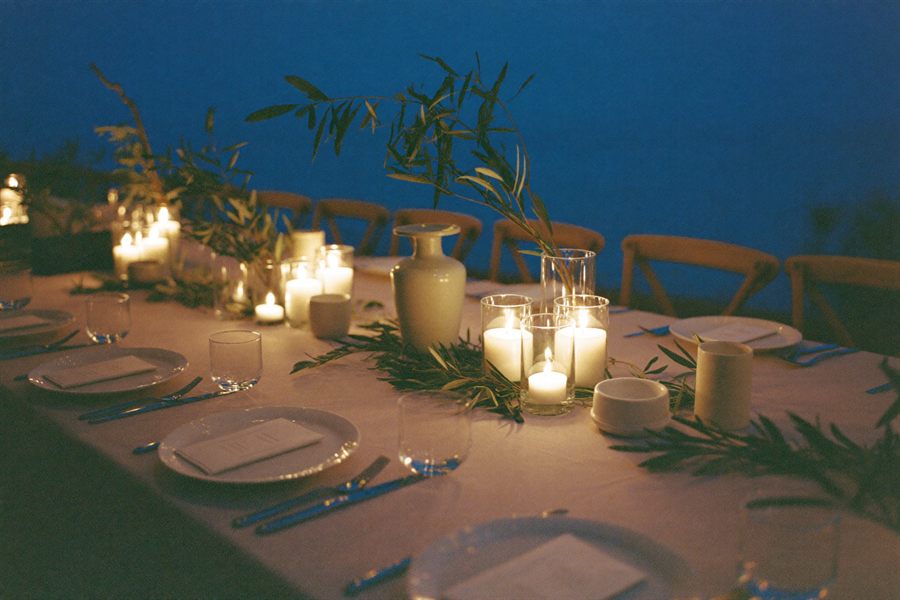 A long linen dinner table set with candles and olive branches at dusk.