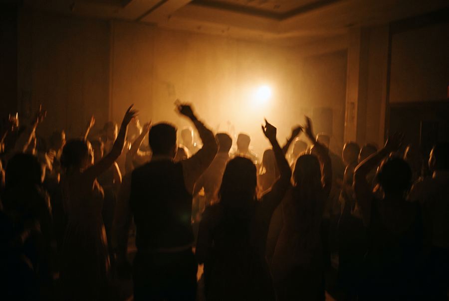 Wedding guests dancing in silhouette with arms raised, blurred with movement and backlit by a single warm light.