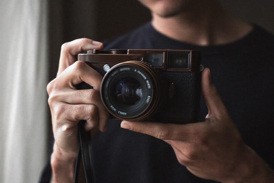 A photographer holding a 35mm film camera at chest height in soft window light.