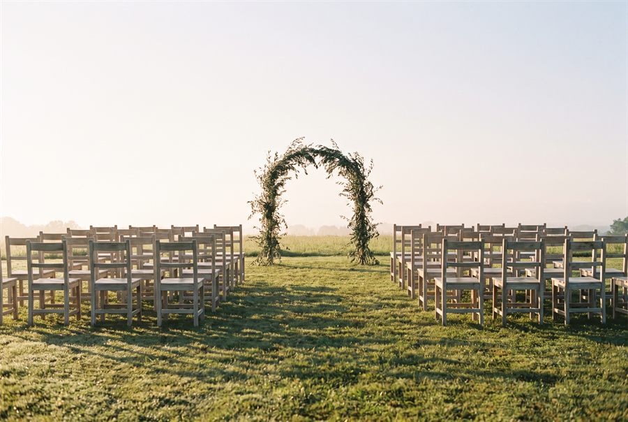 Rows of pale wooden chairs on grass facing an arch of olive branches, before guests arrive.