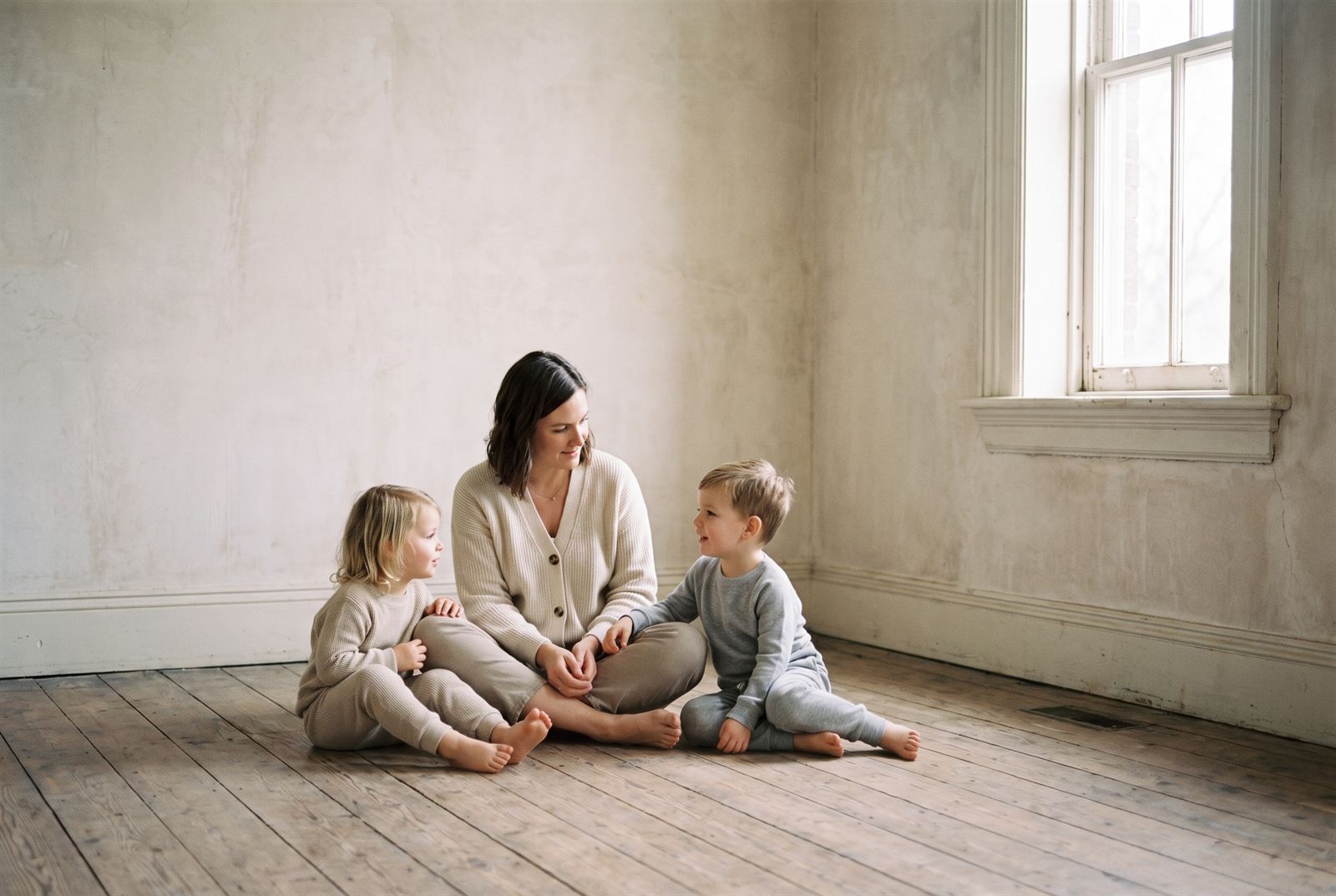 A mother and two young children sitting together on a bare wooden floor in soft window light.