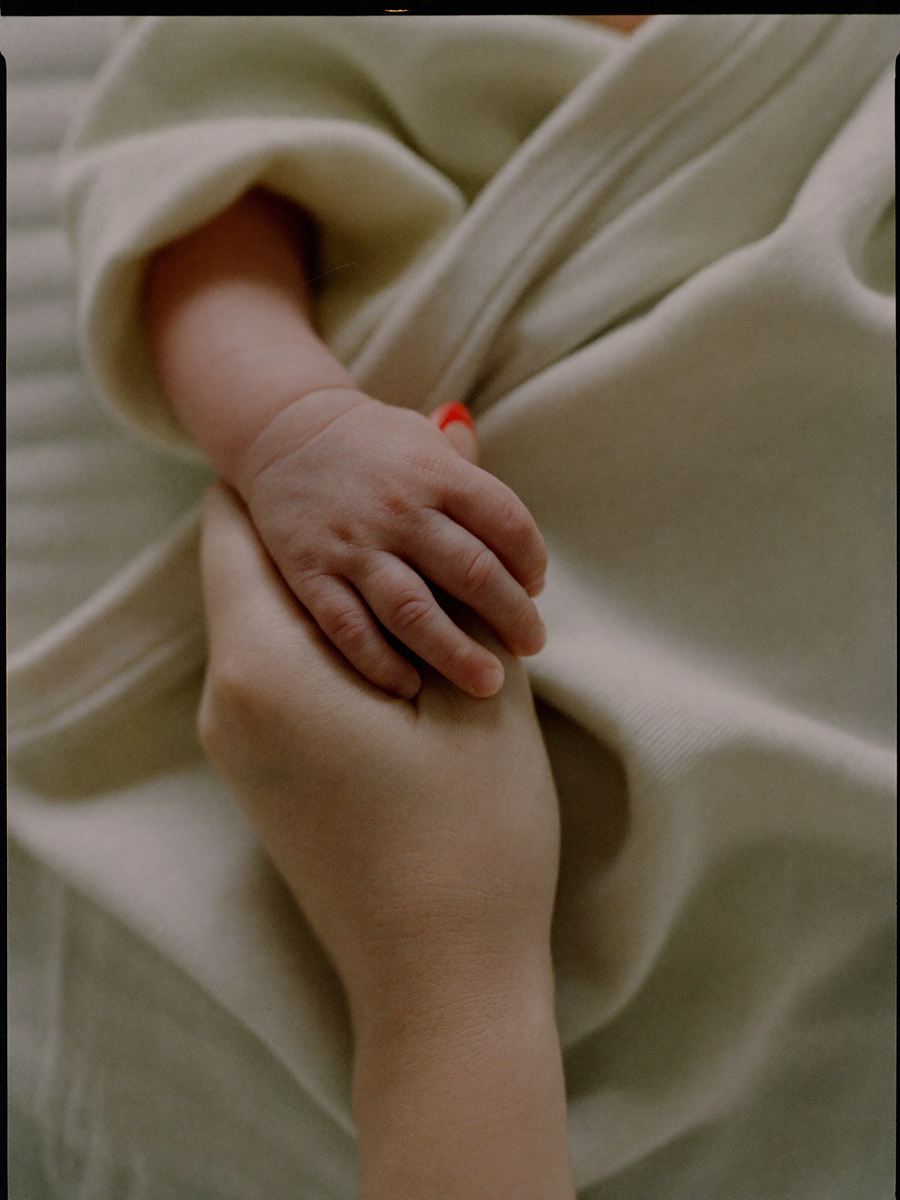 An adult hand cradling a newborn baby's hand against a cream muslin wrap.