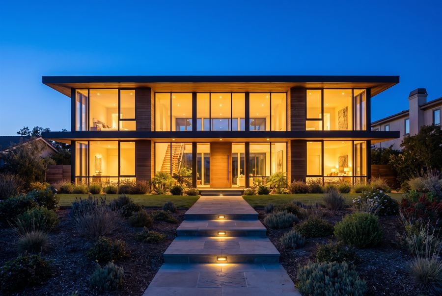 A contemporary house photographed at dusk with every interior light on against a deep blue sky.